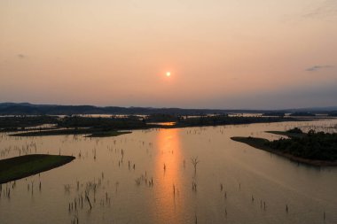 Yumuşak altın ışık, gün batımında Nakai Gölü 'nün sakin sularında dağılmış ağaç kütüklerini ve alçak adaları aydınlatır. Geniş su baskını manzarası açık ve sıcak bir gökyüzünün altındaki uzak tepeler tarafından çerçevelenmiştir..