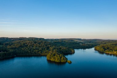 Settons Gölü 'nde sakin mavi sularla çevrili küçük bir ormanlık adanın havadan görünüşü, berrak bir akşam gökyüzünün altında yuvarlanan orman tepeleri..