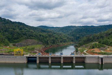 Yoğun yeşil orman ve bulutlu bir gökyüzünün altında yuvarlanan tepelerle çevrili sakin bir su deposunu çevreleyen beton hidroelektrik barajın geniş bir görüntüsü. Sahnede mühendislik ürünü yapılar, yansıtıcı su ve tropikal bir manzaranın doğal dokuları yer alıyor..