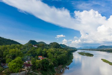 Luang Prabang, Laos yakınlarındaki Mekong Nehri kıyısında geleneksel binalarla dolu yemyeşil tepeler yükseliyor. Ahşap tekneler geniş bir gökyüzünün altında durgun su boyunca uzanır dramatik kümülüs bulutları ve uzak mavi dağlarla dolu..