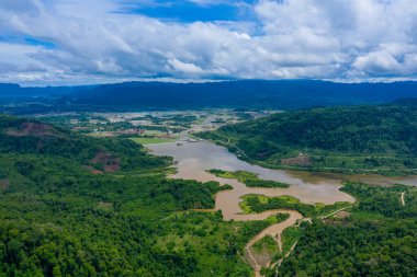 Wide aerial panorama of the Nam Theun 2 Reservoir in central Laos, with winding waterways, lush green hills, and distant mountains under a dynamic sky. The scene highlights irregular shorelines, dense forest, and a mix of natural textures in a