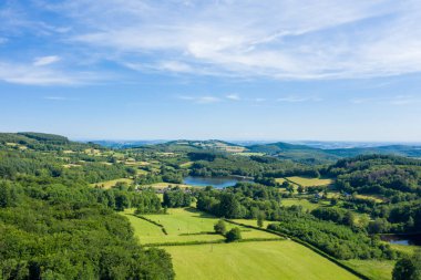 Yuvarlanan yeşil tepeler, yamalı araziler ve Morvan bölgesinde yoğun ormanlık alanlarla çevrili sakin bir göl. Sahne, yumuşak bulutlu, geniş mavi gökyüzünün altında, parlak güneş ışığıyla yıkanır..