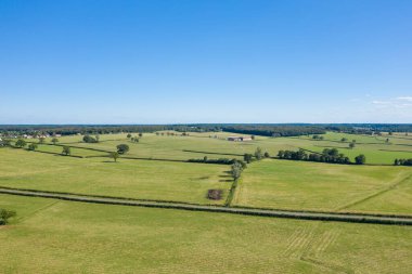 Nivernais kırsalında açık mavi bir gökyüzü altında yeşil çitlere bölünmüş ve ağaçlarla benekli geniş hava manzarası. Manzara geniş, parlak ve sakin, uzak çiftlik binaları ve sakin kırsal bir atmosferi var..