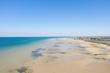 Bernieres-sur-Mer 'deki Juno Plajı' ndaki geniş kumlu kıyı şeridi, turkuaz deniz manzaralı, parlak mavi gökyüzü altında sakin bir sahil köyüyle buluşuyor..