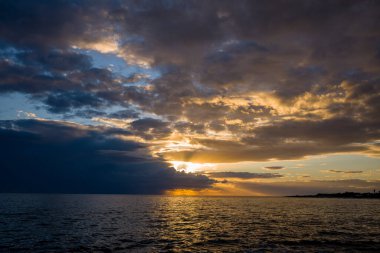 Adriyatik Denizi 'nin yukarısındaki dramatik bulutların içinden geçen ılık güneş ışığı dalgalanan suyun üzerinde altın yansımalar oluşturur. Geniş deniz manzarası, gölgeli gökyüzü ile gün batımında aydınlanan ufuk arasında çarpıcı bir tezat içerir..