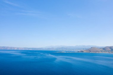Geniş mavi sular ön planda, yumuşak tepeler ve berrak bir gökyüzünün altındaki uzak kıyı şeridinde görülebilen Kissamos limanı ile doludur. Sahne parlak, açık ve sakin..