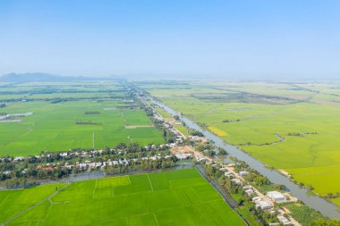 Geniş yeşil pirinç tarlalarından ve Vietnam kırsalındaki küçük bir köyden düz bir kanal geçer. Manzarada geometrik tarım arazileri, dağınık evler ve berrak bir gökyüzünün altındaki uzak puslu dağlar yer alıyor..