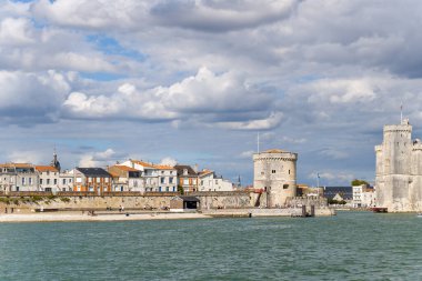 Ortaçağ taş kuleleri ve tarihi rıhtım evleri dramatik katmanlı bulutların altında La Rochelle, Charente-Maritime limanı hizasında. Sahne, güçlendirilmiş mimari, dokulu taş duvarlar ve Atlantik 'in sakin sularını ön plana çıkarıyor.