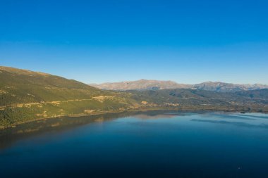 Ioannina Gölü 'nün geniş mavi suları, Yunanistan' ın Epirus kentinde sakin bir manzara yaratan ormanlık yamaçlar ve berrak gökyüzü ile engebeli Pindus dağlarının altında uzanır. Sahne, doğal yansımaları, geniş açık alanı ve açık gün ışığını ön plana çıkarıyor..