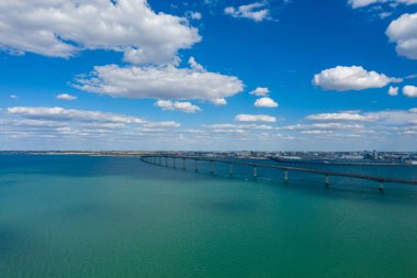 A sweeping view of the Ile de Re bridge extends across clear turquoise water beneath a sky filled with scattered cumulus clouds. The scene captures the open seascape and coastal atmosphere of Charente-Maritime in western France.