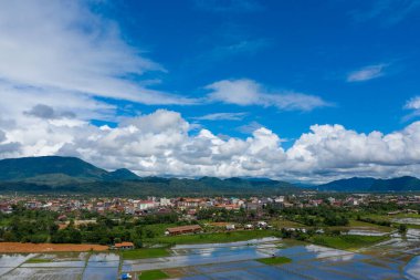 Geniş bir manzara Laos 'un Vang Vieng kasabasını gösteriyor, ön planda su basmış pirinç tarlaları olan yemyeşil dağların eteklerine kurulmuş. Kabarık beyaz bulutlar canlı mavi bir gökyüzünde sürükleniyor, yağmur mevsiminde canlı manzarayı vurguluyor..