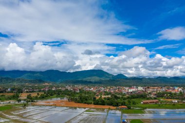 Laos 'un Vang Vieng kasabasının ön planında sel basmış pirinç tarlaları ve dramatik bulutlarla dolu canlı mavi gökyüzünün altında yemyeşil dağlarla dolu panoramik bir manzara. Sahne yağmur mevsiminde kırsal vadiyi vurgular.