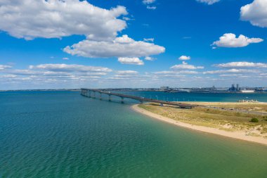 Ile de Re köprüsünün yükseltilmiş manzarası turkuaz suları ve kumlu kıyı şeridini kümülüs bulutlarıyla kaplı bir gökyüzünün altında. Sahne, Batı Fransa 'nın açık kıyı manzarası ve sakin atmosferini vurguluyor..