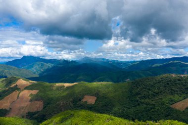 Güneş ışığı demetleri, Phou Khoun ve Luang Prabang, Laos arasındaki dağlık bölgelerdeki dramatik ve dokulu bulutların altında yuvarlanan yeşil tepeleri ve uzak mavi tepeleri aydınlatır. Manzara verimli bitki örtüsü, açık yamaçlar ve canlı bir etkileşim içerir.