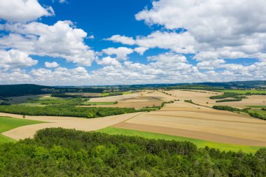 Yoğun ormanlık ve dağınık kümülüs bulutlarıyla dolu bir gökyüzünün altındaki yuvarlanan tepelerle çevrili çeşitli yeşil ve altın tonlarında ekilmiş tarlaların geniş hava manzarası. Manzara bir açıklık ve kırsal sakinlik hissi yayıyor..