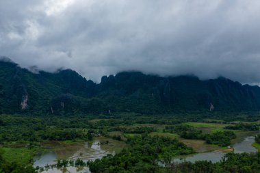 Alçak bulutlar, sel basmış pirinç tarlaları ve Laos 'un Vang Vieng yakınlarındaki yoğun tropikal ormanın üzerindeki dramatik kireçtaşı tepeleri boyunca sürükleniyor. Karamsar atmosfer, engebeli uçurumlar ve yemyeşil manzaralar arasındaki zıtlığı vurgular..