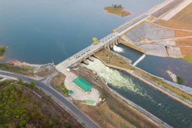 Geniş hava perspektifi, sakin bir su deposunu çevreleyen beton bir hidroelektrik barajını ve kontrol edilen bir kanala akan suyu gösteriyor. Çevresindeki arazide mühendislik harikası setler, erişim yolları ve altında seyrek bitki örtüsü bulunur.