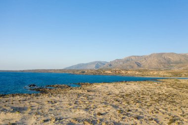 Geniş kumlu kıyı şeridi ve seyrek bitki örtüsü, Girit 'teki Elafonisi' de berrak mavi gökyüzünün altında turkuaz suyla ve engebeli dağlarla buluşuyor. Sahne güneşli ve huzurlu, doğal dokuları ve kıyı güzelliğini vurguluyor..