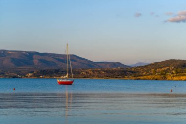 Canlı kırmızı bir yelkenli sakin mavi sularda süzülür, yumuşak tepeler ve uzak dağlarla çevrili, yumuşak altın bir akşam gökyüzünün altında. Sakin sahne ince yansımalar ve sıcak doğal ışık içerir..