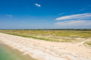 Quiberon 'daki Plage du Mentor' un geniş kumlu bir kıyı şeridi, yumuşak sörf ve canlı mavi gökyüzü altında yuvarlanan çimenli kum tepelerinin geniş bir görüntüsü. Manzara açık, güneşli ve doğal dokular ve yumuşak renklerle huzur dolu..