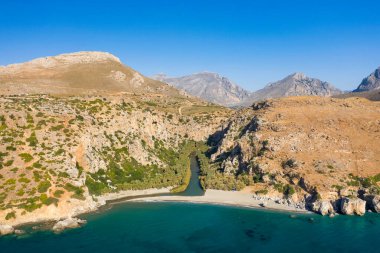 Engebeli bir dağ vadisinde süzülen dar bir nehrin ve Girit, Preveli 'deki boş kumlu plajın hava görüntüsü. Manzara kurak kayalık yamaçlar, seyrek yeşillik ve bulutsuz bir gökyüzünün altında derin mavi deniz içerir..