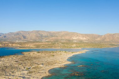 Yunanistan 'ın Girit kentindeki Elafonisi sahilinde şeffaf turkuaz suyla seyrek bitki örtüsü buluşan kumlu bir yarımadanın hava manzarası bulutsuz bir gökyüzü altında engebeli, güneşli dağlar tarafından destekleniyor. Manzara kuru, geniş ve doğal olarak canlı görünüyor..