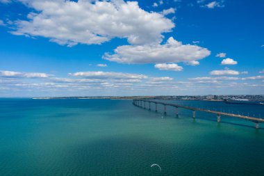 Uzun beton köprü, bulutlarla dolu bir gökyüzünün altında, anakarayı Ile de Re 'ye bağlayan, berrak turkuaz suyun üzerinde yumuşak bir yay şeklinde uzanır. Geniş deniz manzarası açık kıyı ortamını ve sakin atmosferi vurguluyor