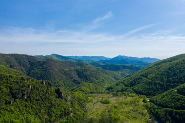 Foret domaniale de la Vis 'deki berrak mavi gökyüzünün altındaki yemyeşil tepelerin ve sık ormanın havadan görünüşü. Güneş ışığı kayan araziyi ve derin vadileri vurguluyor, bir sükunet ve enginlik hissi yaratıyor..