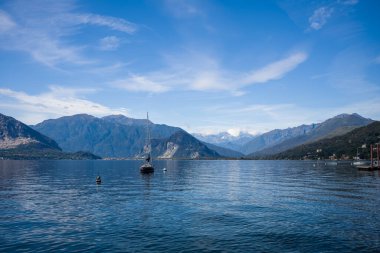 Kuzey İtalya 'daki Maggiore Gölü' nün sakin mavi sularında, ormanlık tepeler ve yumuşak bulutlarla kaplı gökyüzünün altındaki uzak dağlarla çevrili bir yelkenli yüzer. Huzurlu sahne doğal güzelliği ve huzuru vurguluyor.