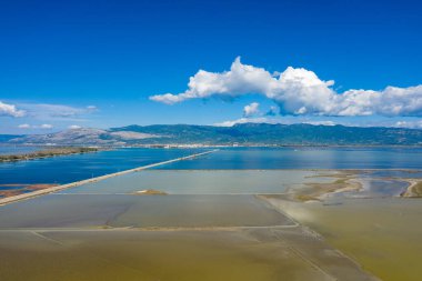 Uzun düz bir yol, Yunanistan 'ın batısındaki Missolonghi' deki geometrik tuz bataklıklarını parlak bir gökyüzü altında canlı mavi sular ve uzak dağlarla böler. Geniş sulak arazide beyaz bulutlar sürükleniyor, benzersiz manzarayı ve sükuneti vurguluyor.