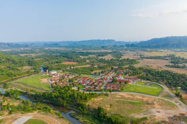 Yeşil tarlalar, dolambaçlı yollar ve kıvrımlı nehirlerle çevrili kırmızı çatılı evleri olan modern bir kırsal yerleşim alanı. Manzarada açık kırsal alanlar, dağınık ormanlar ve açık gün ışığı altında uzak sıradağlar yer almaktadır..