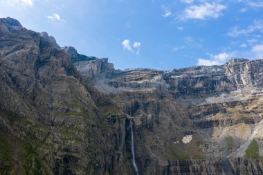 Cirque de Gavarnie 'de büyük katmanlı kaya oluşumları dramatik bir şekilde yükseliyor. İnce bir şelale, engebeli uçurumlardan dökülüyor. Parlak mavi gökyüzü ve dağınık bulutlar altında..