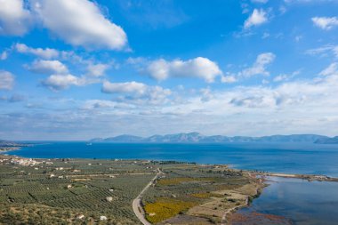 Tarımsal tarlalarla çevrili geniş bir kıyı şeridi, yansıtıcı bir lagün ve dağınık bulutlarla dolu canlı mavi bir gökyüzünün altında uzak sıradağları barındıran hava manzarası. Sahne açıklık ve doğal güzelliği dokuların karışımı ve...