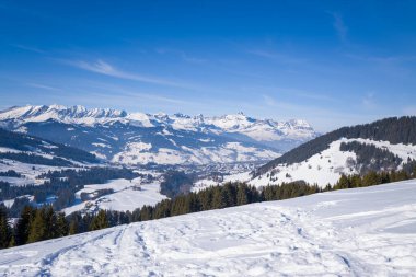 Karla kaplı tepeler ve Megeve köyü aşağıdaki vadide yuva yapmış, dramatik alp zirveleriyle çevrelenmiş, canlı mavi gökyüzü altında sonsuz yeşil ormanlar. Parlak güneş ışığı kışın serin dokularını ve geniş manzarasını güçlendirir..