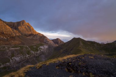 Sıcak bir akşam güneşi, engebeli dağ kayalıklarına ve Pireneler 'deki çimenli yamaçlara çarpıcı gölgeler ve renkli bir gökyüzüne dokunur. Huzurlu, doğal bir manzara yaratır..
