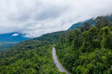 Kıvrımlı bir yol, yoğun yeşil tropikal ormanları kesiyor. Dağlık arazide alçak bulutlar asılı. Sahne, Laos 'un kırsal kesimlerinde yavaş seyahat ve macera çağrıştırıyor..