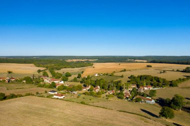 Geniş altın tarlalar ve dağınık ağaçlar arasında yuva yapmış, Fransa 'nın orta kesimindeki canlı mavi gökyüzünün altında ağaçlarla çevrili Cuncy les Varzy köyünün yüksek açılı manzarası. Açık arazi ve yumuşak tepeler kırsal sakinlik ve ferahlık hissi yaratıyor..