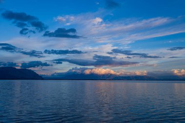 Sıcak günbatımı ışıklarıyla aydınlanan katmanlı bulutlar uzak Mora dağları üzerinde sürüklenerek geniş bir gölün sakin yüzeyine hafifçe yansıyor. Huzurlu manzara yumuşak dalgalanmalar, geniş gökyüzü dokuları ve ince pastel renkler içerir..