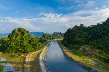 Dik, orman kireçtaşından tepeler arasında düz bir kanal uzanır. Canlı, mavi bir gökyüzünün altında dağınık bulutlarla. Manzara verimli yeşillik, kayalık yamaçlar ve yansıtıcı su içerir ve mühendislik yapısı ile mühendislik arasında çarpıcı bir kontrast oluşturur.