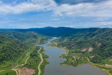 Yoğun ormanlık tepeler ve parlak bir gökyüzünün altındaki temiz araziler arasında dolaşan bir nehrin havadan perspektifi. Çevrede dolambaçlı su, yeşil yamaçlar ve uzak kırsal bölgelerde doğal eğriler bulunur..