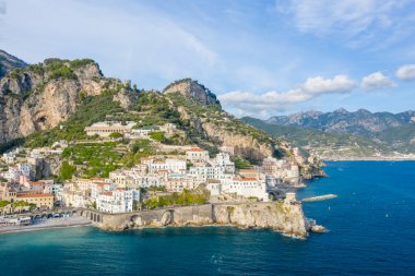 A wide aerial perspective captures the sunlit whitewashed buildings of Amalfi, Italy, cascading down rugged cliffs toward the vivid blue Mediterranean Sea. The dramatic shoreline features historic stone arches, lush greenery, and a mountainous