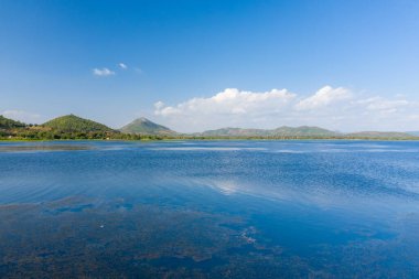 Brateak Gölü 'nün derin mavi suları Kamboçya' nın Kampong Chhnang şehrinde yeşil bir kıyı şeridine ve hafif eğimli tepelere doğru uzanır. İnce su bitkileri ve dağınık bulutlar geniş ve sakin manzaraya dokular ekler..