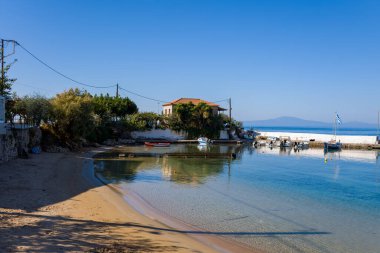 Berrak mavi suyu ve küçük tekneleri olan sakin bir liman kumlu bir sahil kenarında ve bulutsuz bir gökyüzünün altında geleneksel bir evdir. Sakin yansımalar ve yemyeşil yeşil huzurlu bir Akdeniz kıyısı manzarası yaratır.