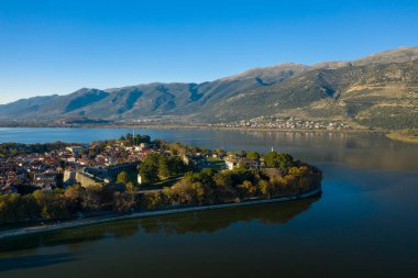 Arkaplanda engebeli Pindus dağları yükselirken, Ioannina Gölü ile çevrili tarihi eski Ioannina kasabasını geniş bir hava manzarası yakalıyor. Görüntü ortaçağ duvarlarını, kırmızı kiremitli çatıları ve berrak mavi gökyüzünün altındaki sakin suyu ön plana çıkarıyor.