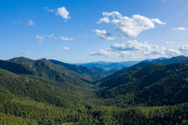 İtalya, Sardunya 'da engebeli dağlar ve sık ağaçlıklarla çevrili engin yeşil bir vadinin havadan görünüşü. Kabarık bulutlar berrak mavi bir gökyüzünde sürükleniyor, manzaranın üzerine yumuşak gölgeler bırakıyor..