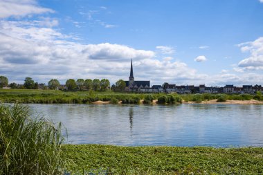 Geleneksel köy binaları ve yüksek bir kilise çan kulesi ufukta, parlak mavi gökyüzünün altında yemyeşil kıyıları olan Loire nehrinin sakin sularında yansıtılıyor..