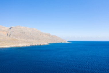 Girit 'in Kato Zakros kenti yakınlarındaki engebeli ve güneşli bir kayalık sahil şeridine ulaşan derin mavi Akdeniz sularının geniş manzarası. Sahne berrak gökyüzü, minimum bitki örtüsü ve sakin, açık bir atmosfer ile tanımlanır..