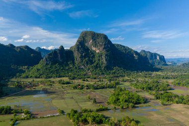 Su basmış pirinç tarlaları ve bereketli kırsal bölgelerdeki dağınık kır evleri mozaiğinin üzerinde dramatik kireçtaşı kayalıkları yükseliyor. Canlı mavi gökyüzü ve canlı yeşillik, Güneydoğu Asya kırsalının tipik doğal desenlerini ve açık alanını vurguluyor.