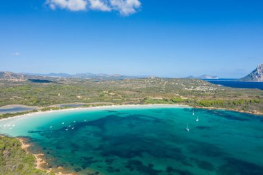 Sardinya 'daki Cala Brandinchi plajının yüksek açılı görüntüsü turkuvaz suyu, demirli tekneler ve uzak dağlarla dolu açık bir gökyüzünün altındaki yemyeşil kıyı şeridini gösteriyor..