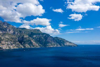 Dik yeşil kayalıklar, dağınık beyaz bulutlarla benekli bir gökyüzünün altındaki derin mavi Akdeniz sularına inerler. Sakin sahne, İtalyan kırsalının dramatik manzarasını ve canlı kıyı renklerini vurguluyor..